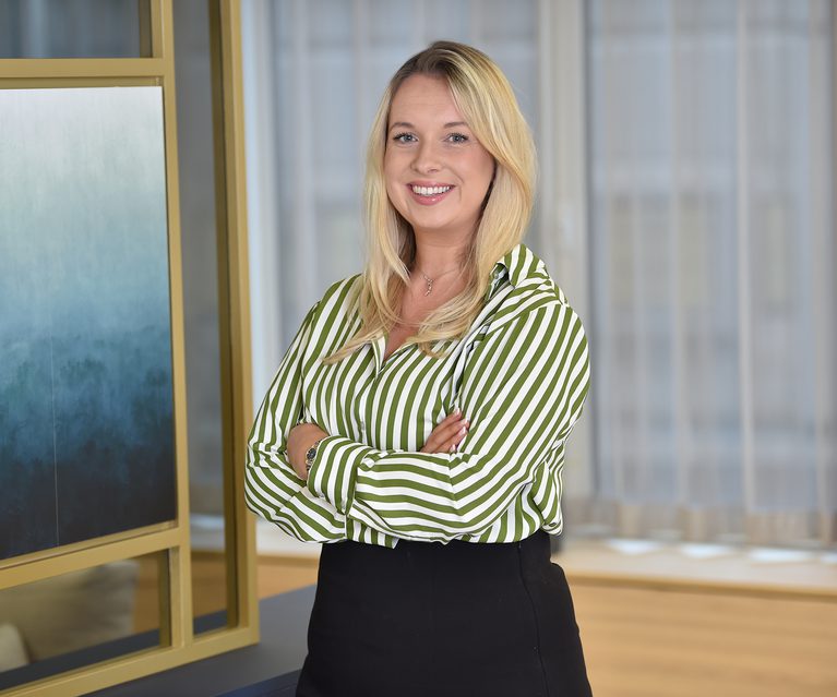 A smiling blonde woman with crossed arms stands in a modern office with glass partitions and a wooden floor, wearing a green and white striped shirt and a black skirt.