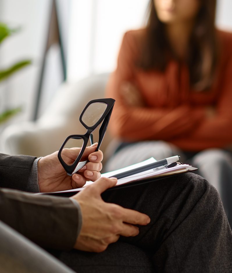 Close-up of a person in dark clothing holding black glasses and a clipboard, with two blurred individuals seated on a sofa in a bright living room.
