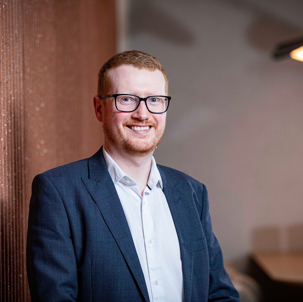 Portrait of a person with short red hair and glasses, wearing a navy blazer and white shirt, smiling while leaning against a brick wall.