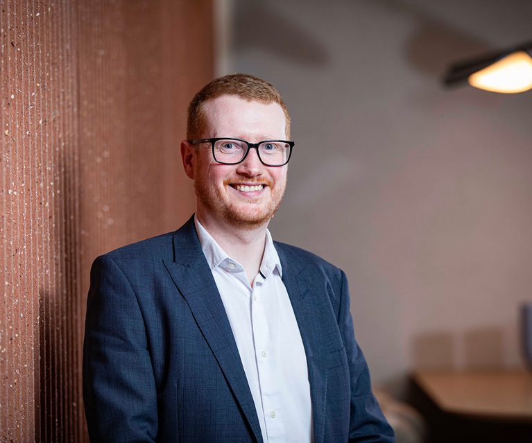 Portrait of a person with short red hair and glasses, wearing a navy blazer and white shirt, smiling while leaning against a brick wall.