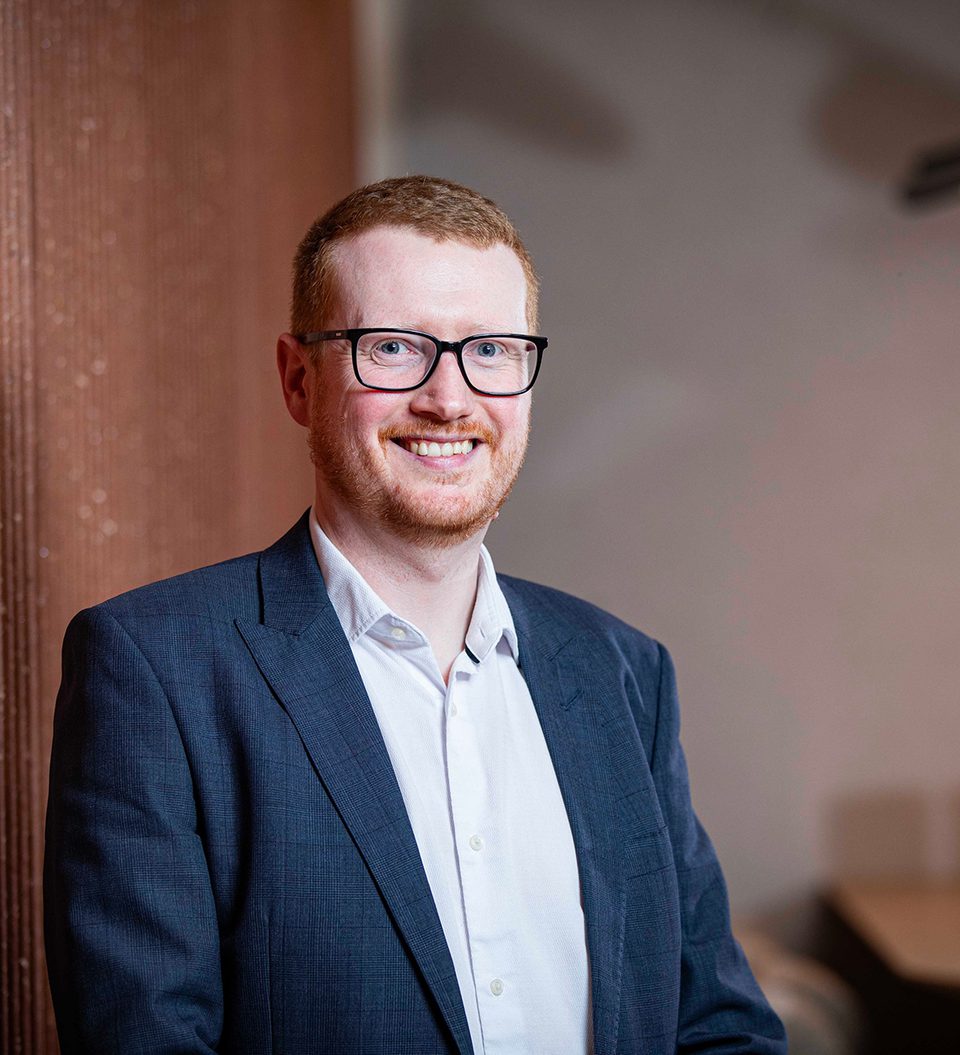 Portrait of a person with short red hair and glasses, wearing a navy blazer and white shirt, smiling while leaning against a brick wall.