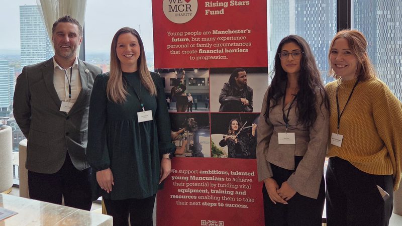 Four people standing indoors with a cityscape visible through large windows behind them, next to a red and white display board promoting Manchester's Rising Stars Fund.