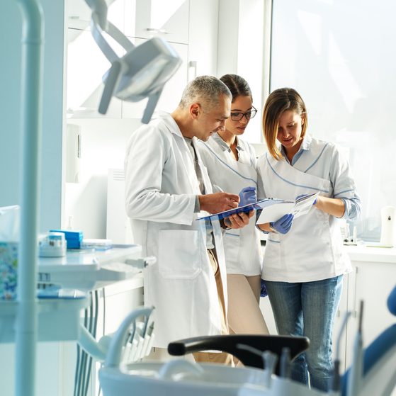 Three healthcare professionals in white coats and gloves discussing documents in a brightly lit dental clinic, with dental equipment and a chair visible.