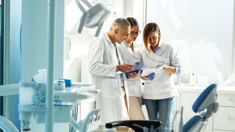 Three healthcare professionals in white coats and gloves discussing documents in a brightly lit dental clinic, with dental equipment and a chair visible.