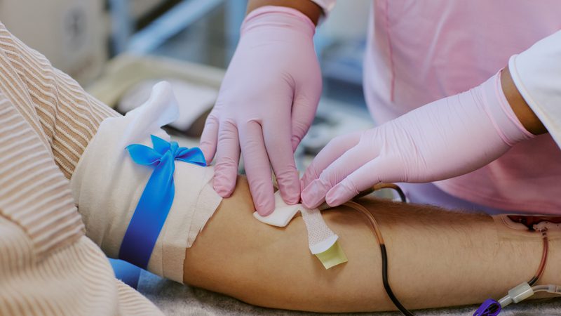 A healthcare professional in pink scrubs and pink gloves is inserting a needle into a patient's arm during a medical procedure, with the patient's arm taped and prepared.