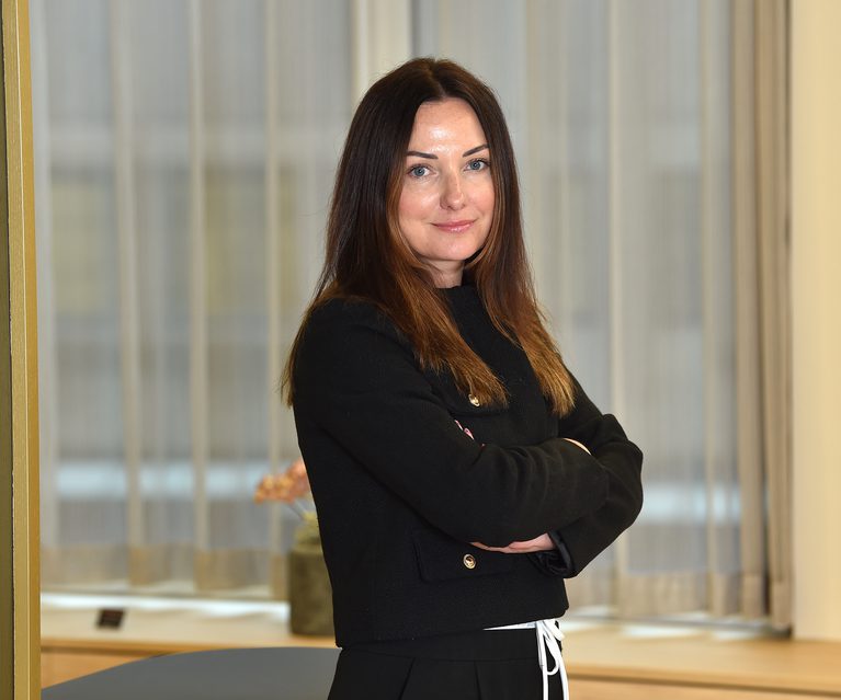 A woman with long brown hair, wearing a black top, stands with her arms crossed in front of a window with beige curtains, smiling gently.