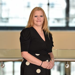 A woman with long blonde hair, wearing a black top with puffed sleeves and a black skirt with large white polka dots, is standing indoors by a metal handrail.
