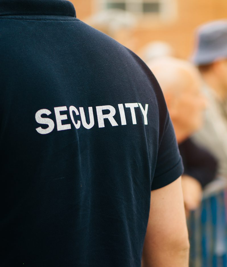 A security guard in a navy blue shirt with "SECURITY" written on the back, standing in front of an outdoor crowd behind a blue barrier.