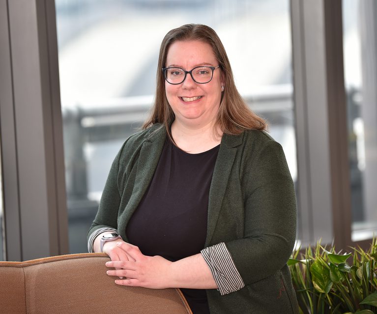 A woman with glasses, long brown hair, and a friendly smile, wearing a black top and dark green blazer, standing indoors near windows with greenery.