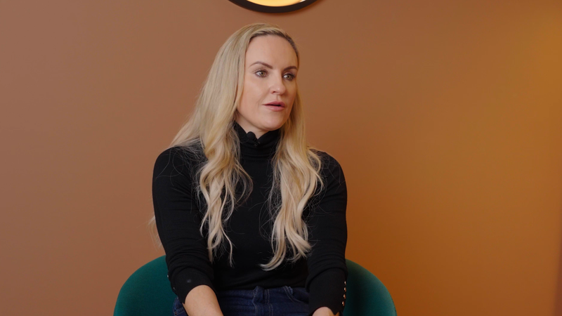 A blonde woman with long wavy hair is seated against a warm brown wall, wearing a black turtleneck with buttoned sleeves and dark jeans.