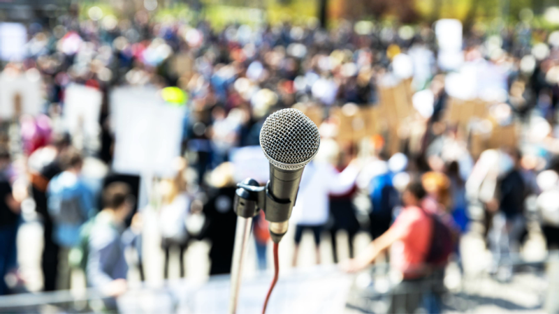 A close-up of a microphone in front of a blurred crowd at an outdoor event or protest.