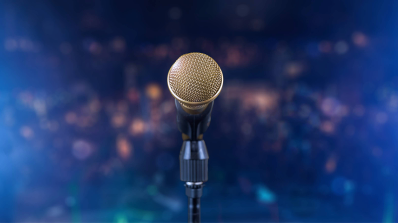 Close-up of a gold mesh microphone on a stand, centered on a stage with vibrant blue bokeh lights in the background, suggesting a live performance setting.