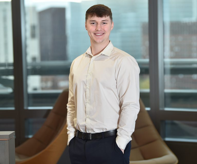 Smiling person wearing a light beige shirt and dark trousers stands with hands in pockets in a modern office with large windows.