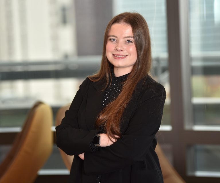 A young woman with long, straight, brown hair, wearing a black blazer and a polka-dot top, standing with arms crossed in an office setting with large windows.