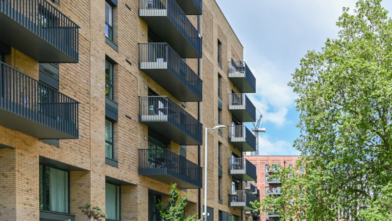 A multi-storey residential building with brick walls and black balconies, with a tree on the right and a partly cloudy sky overhead.