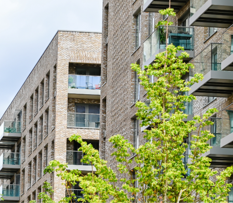 Modern multi-storey apartment building with brick facade, glass balconies, and a leafy tree in the foreground under a partly cloudy sky.