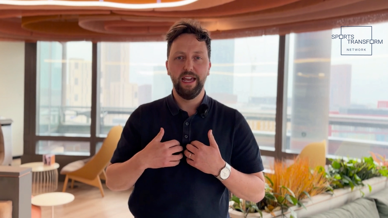 Bearded person in a dark polo speaks to camera, hands on chest, in a bright modern office with large windows and plants; Sports Transform Network logo in the corner.