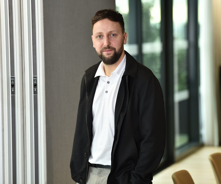 A man with short dark hair and a beard, wearing a white polo shirt and black jacket, standing indoors with glass doors and greenery in the background.