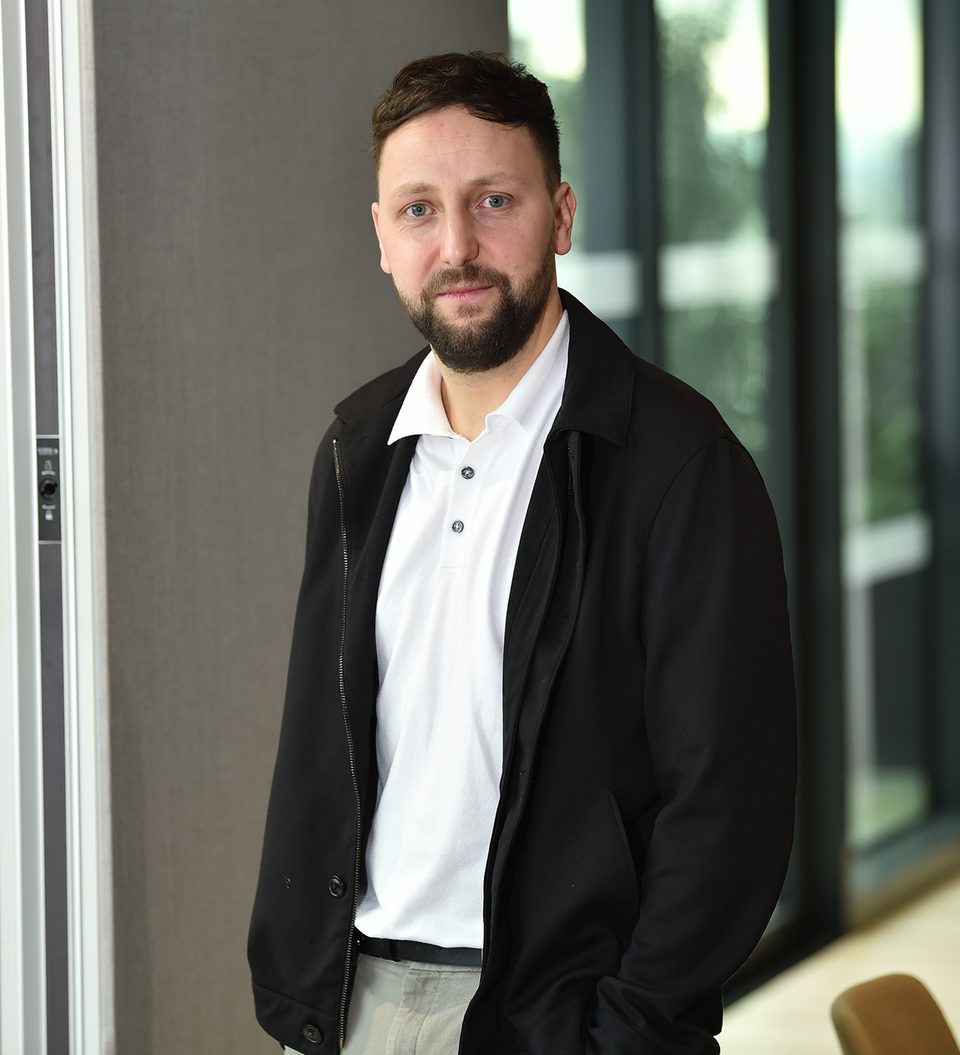A man with short dark hair and a beard, wearing a white polo shirt and black jacket, standing indoors with glass doors and greenery in the background.