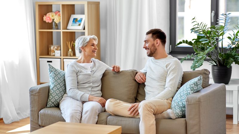 An elderly woman and a young man are sitting on a beige sofa, smiling and enjoying a conversation in a bright living room with a window, bookshelf, and houseplant.