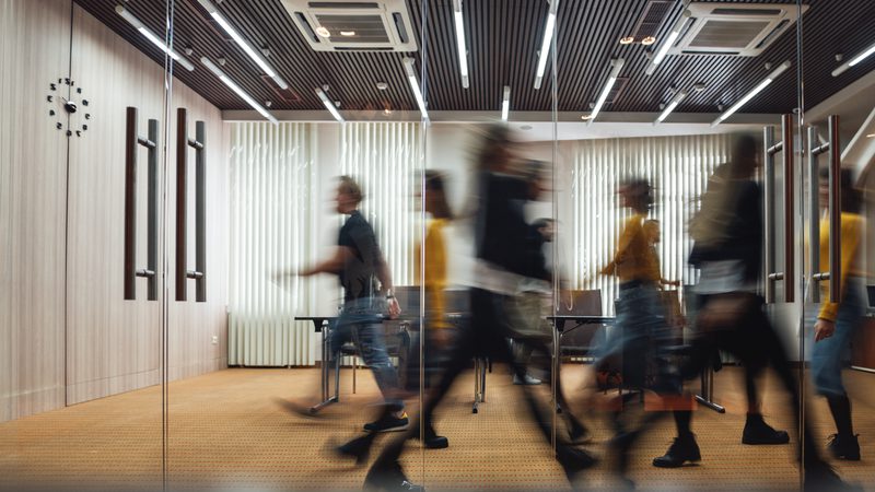 Blurry figures walking in a modern conference room with mirrored glass wall, wood paneling, vertical blinds, and a clock on the wall.