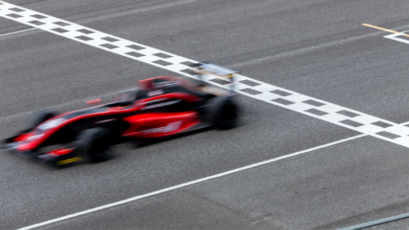 A red Formula 1‑style single‑seater car on a grey race track, captured in motion blur as it speeds past a diagonal black‑and‑white chequered finish line.