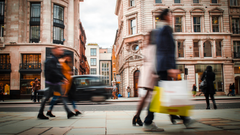 Blurry pedestrians cross a busy city street past pink stone buildings; a moving vehicle streaks through the scene while shoppers carry bags.