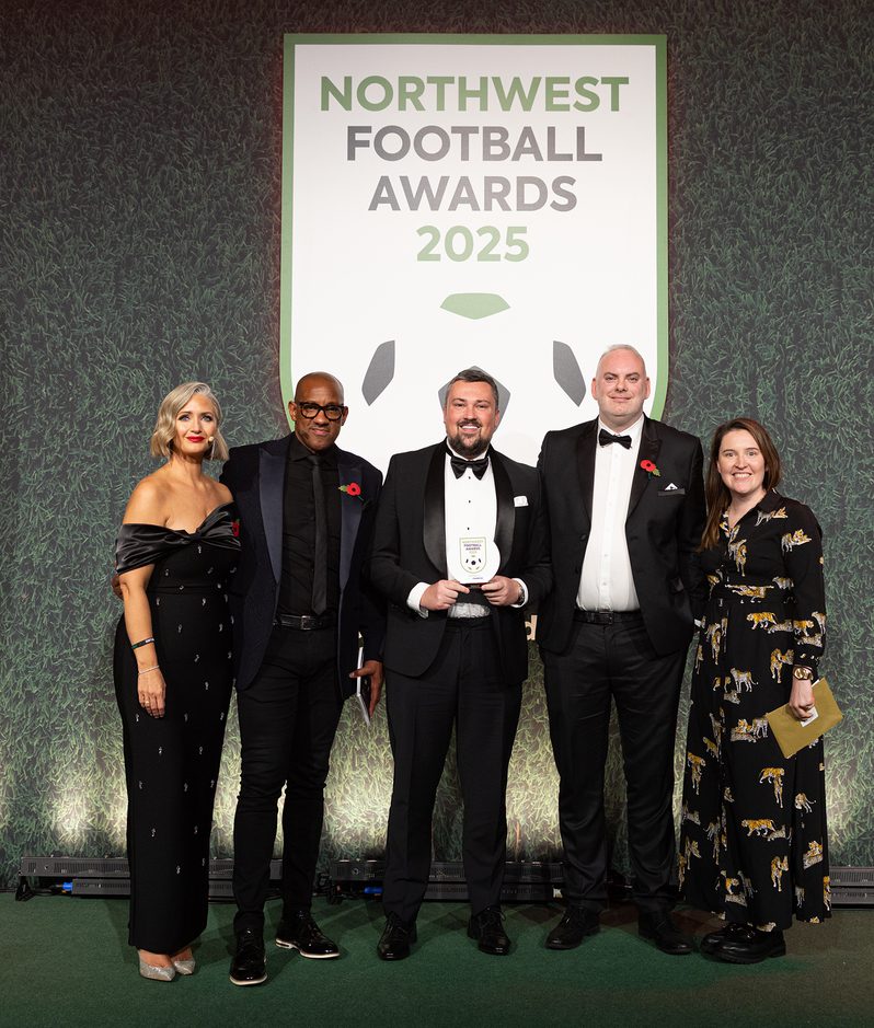 Five people in formal attire pose on stage at the Northwest Football Awards 2025, with a central trophy and a large sign against a textured green backdrop.