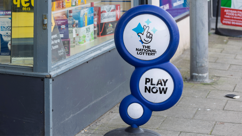 Blue circular sign featuring The National Lottery logo and Play Now text, mounted on a blue stand beside a glass-fronted shop window on a grey pavement.