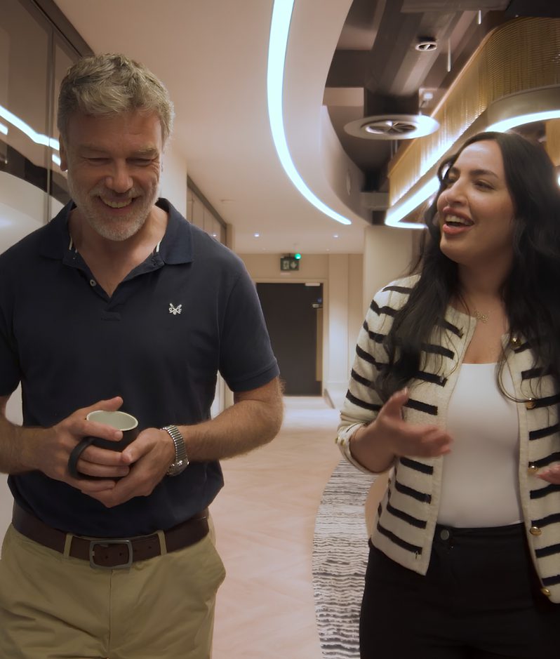 Two people walk and chat in a modern office lounge with curved neon lights, wooden shelves and potted plants, one holding a cup and the other gesturing.