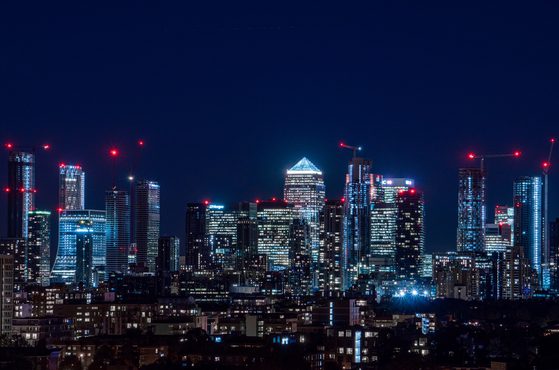 Nighttime city skyline with illuminated skyscrapers, some under construction with red cranes, against a dark sky.