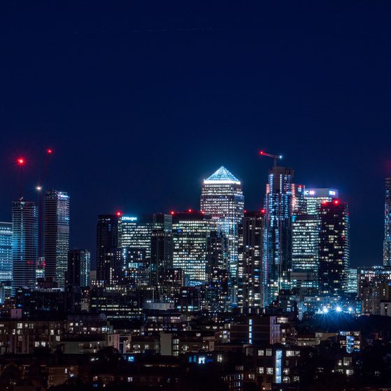 Nighttime city skyline with illuminated skyscrapers, some under construction with red cranes, against a dark sky.
