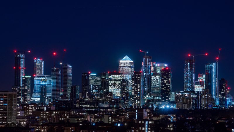 Nighttime city skyline with illuminated skyscrapers, some under construction with red cranes, against a dark sky.