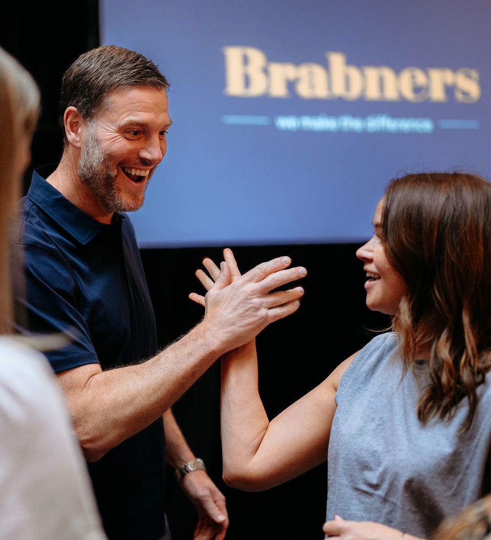 A man and woman smiling and high-fiving each other at an indoor event, with a blurred woman in the foreground and a "Brabners" logo on a screen in the background.