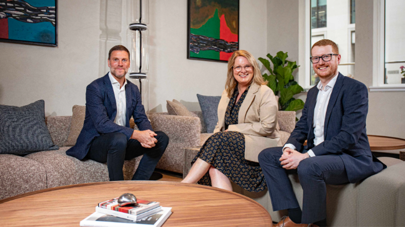 Three professionally dressed individuals sit on a grey sofa in a bright office, smiling at the camera, with abstract wall art and a coffee table with magazines.
