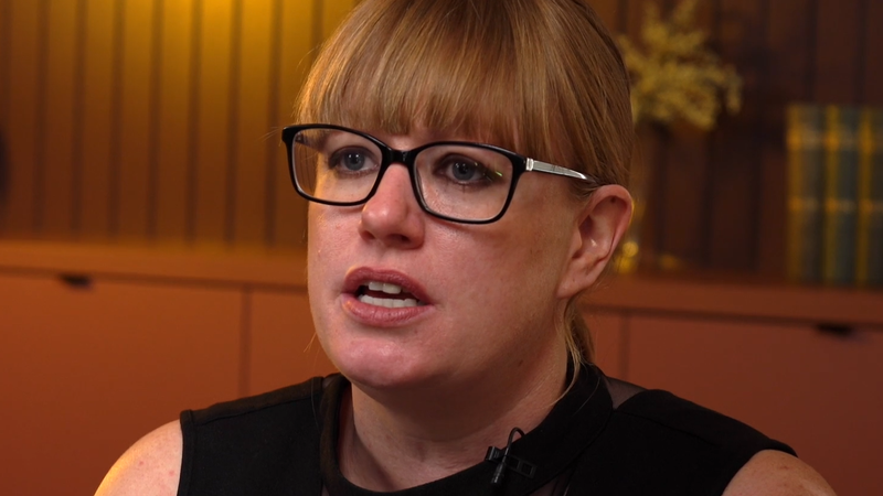 A woman with red hair, wearing black glasses and a sleeveless black top, speaking in a room with wood-paneled walls and a blurred background.