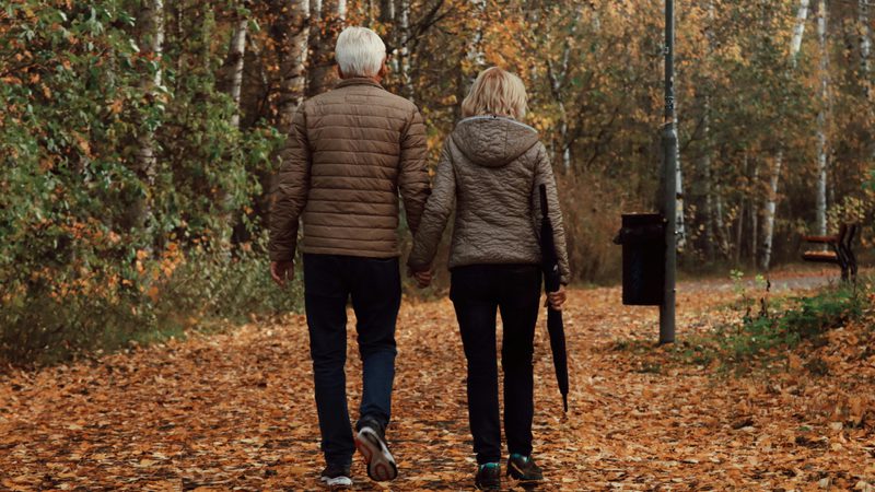 A senior couple walking hand-in-hand along a leaf-covered path in a forest during autumn, surrounded by trees with orange and yellow foliage.