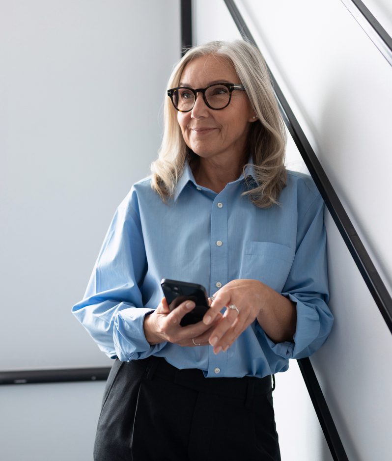 An adult with shoulder-length grey hair and black-framed glasses, wearing a light blue shirt and dark trousers, holds a smartphone beside a bright window.