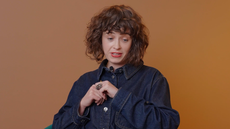 A woman with curly brown hair is wearing a denim jacket and holding her hands together, with a neutral expression against an orange background.