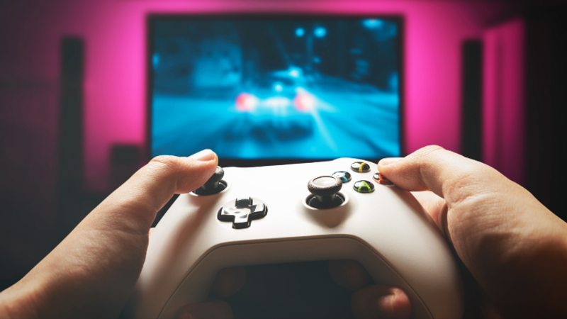 Hands grip a white game controller in the foreground as a blue racing game runs on a screen, with pink neon lighting framing the blurred background.