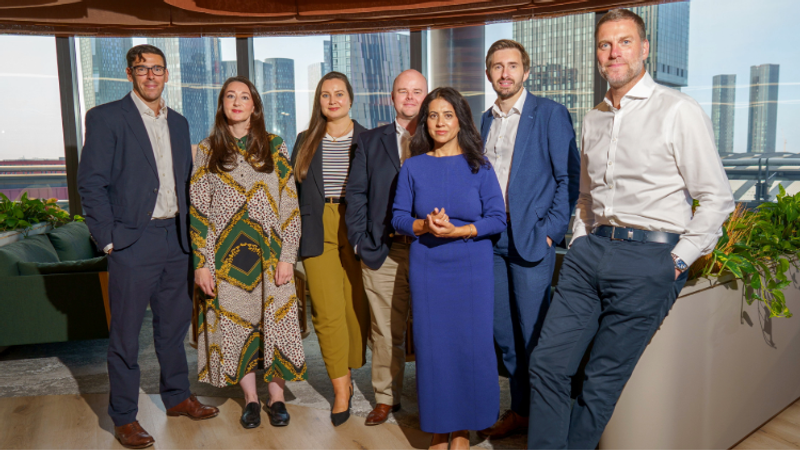 Seven professionals pose in a modern office with floor-to-ceiling windows overlooking a city skyline, dressed in smart business attire and composed expressions.
