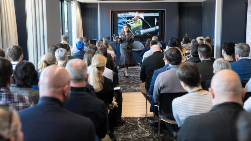 A woman presents on stage in front of a seated audience during a conference or seminar, with a large screen displaying a slide behind her.