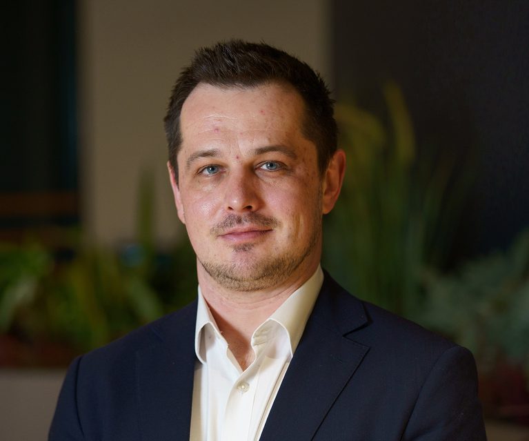 A man with short dark hair, blue eyes, and light facial hair, wearing a dark suit and white shirt, standing indoors with a blurred plant background.