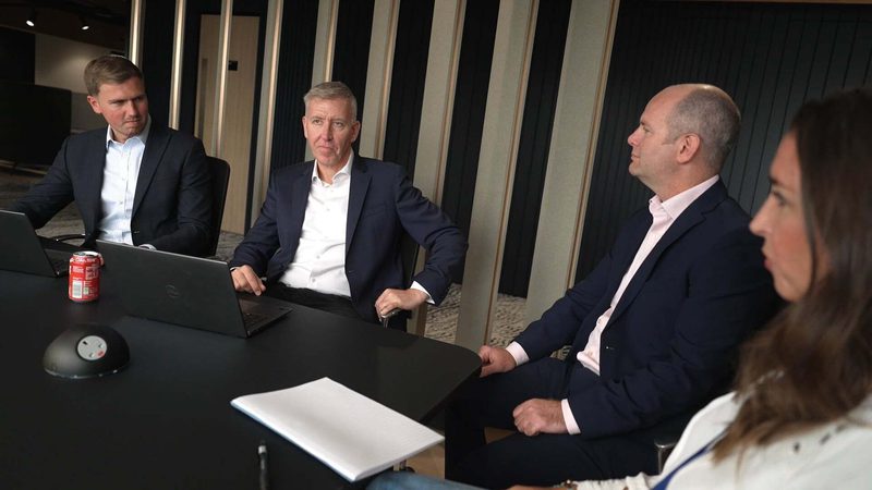 Five professionals seated at a conference table, engaged in a serious meeting; laptops, a soda can, a notepad, and a conference speaker are on the table.