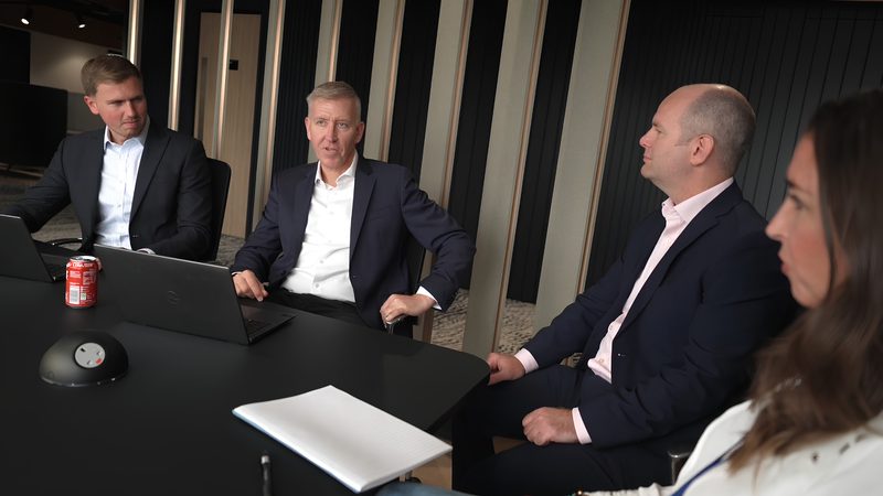 Four people in business attire seated around a black conference table with laptops, a Coca-Cola can, a microphone, and a notebook in a modern office setting.
