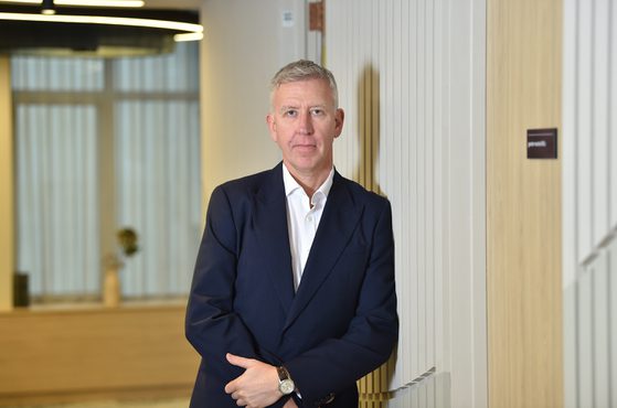 A man in a dark suit with a white shirt, standing with arms crossed against a striped wall in a modern office interior.