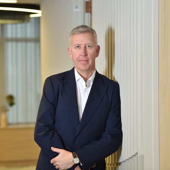 A man in a dark suit with a white shirt, standing with arms crossed against a striped wall in a modern office interior.