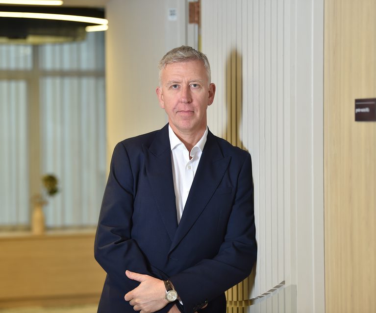A man in a dark suit with a white shirt, standing with arms crossed against a striped wall in a modern office interior.