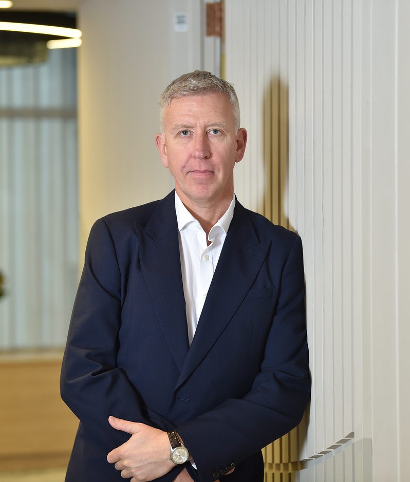 A man in a dark suit with a white shirt, standing with arms crossed against a striped wall in a modern office interior.