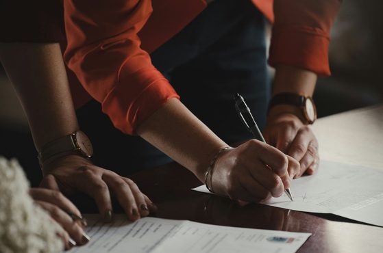 A person in an orange shirt is leaning over a table, signing a document with a pen, with their left hand resting on another document.